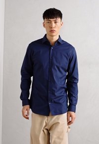 Cămașă albastru marin cu mâneci lungi, cu guler ascuțit și închidere cu nasturi în față, realizată dintr-un material neted. Croială ajustată, cu un design ușor conic.