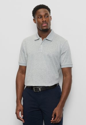 Mann trägt hellgraues Poloshirt und dunkle Hose, steht vor einem schlichten hellgrauen Hintergrund und schaut leicht zur Seite.