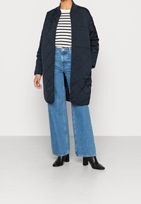 Cappotto lungo trapuntato blu navy con una vestibilità rilassata, caratterizzato da un collo rotondo e tasche laterali. Abbinato a una camicia a righe e jeans di denim a gamba larga.