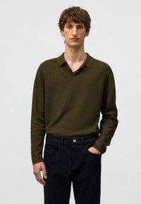 Olivgrünes Langarm-Polohemden mit strukturiertem Muster und Kragen, kombiniert mit schwarzen Hosen. Knöpfe sind am Kragen sichtbar.