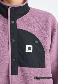 Chaqueta de forro polar púrpura con bolsillo en el pecho negro, botones a presión, bolsillo con cremallera y parche del logotipo de Carhartt.