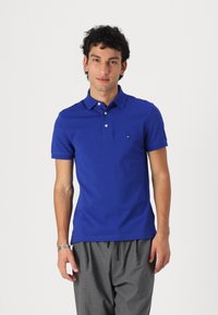 Polo en azul real hecho de algodón, con un collar clásico, mangas cortas y un pequeño logo en el lado izquierdo del pecho. Llevado con pantalones grises.