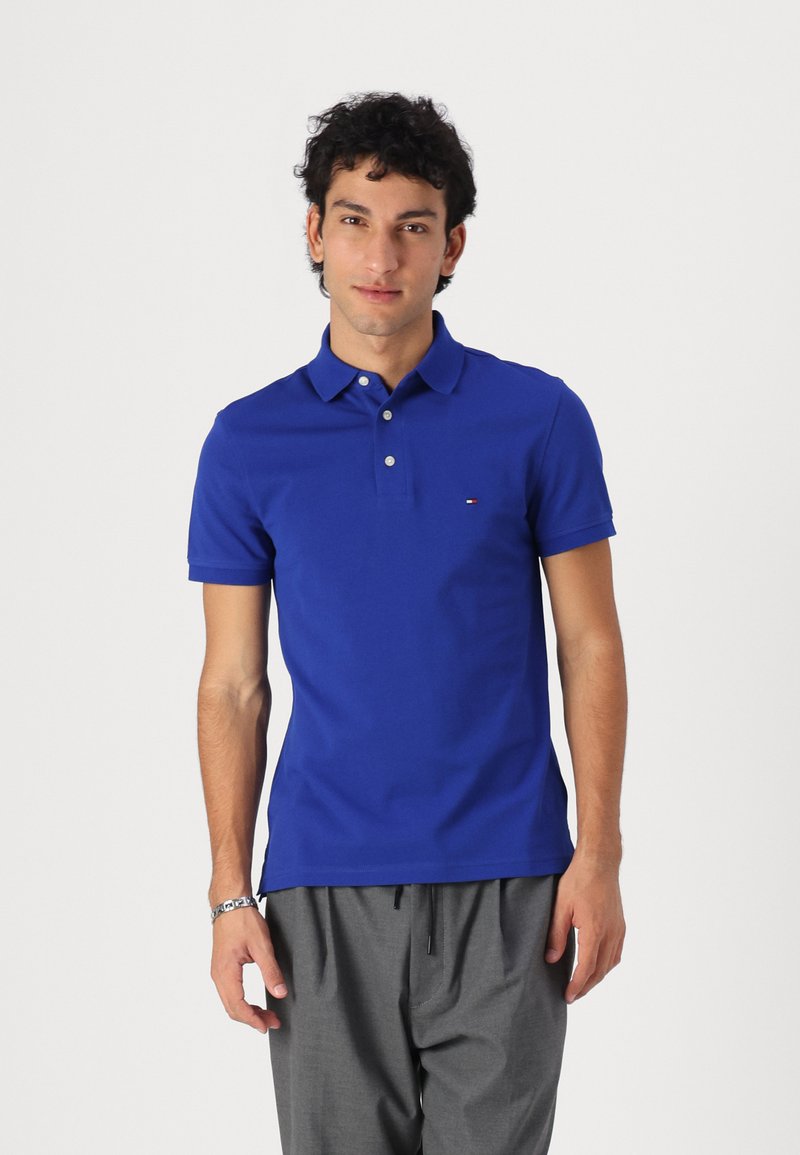 Polo en azul real hecho de algodón, con un collar clásico, mangas cortas y un pequeño logo en el lado izquierdo del pecho. Llevado con pantalones grises.