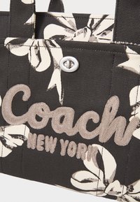 Poche de sac en toile avec imprimé floral beige, texte brodé "Coach New York" en beige, et fermoir tournant argenté.