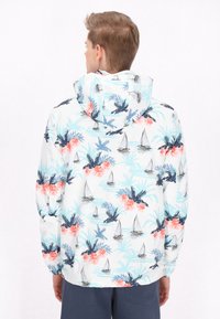 Giacca a vento con base bianca decorata da un motivo tropicale di fiori rosa, foglie di palma e velieri blu e neri. Design con cappuccio.