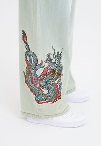 Lichtgroene denim broek met een kleurrijke geborduurde drakendesign op de zijkant, gecombineerd met witte sneakers.