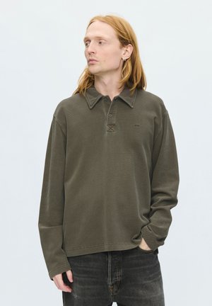 Ung person med langt rødt hår iført olivengrøn langærmet polo-shirt og sorte jeans, kigger sidelæns med den ene hånd i lommen mod ensfarvet baggrund.