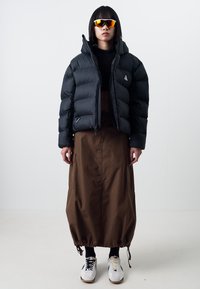 Black puffer jacket with a cropped design, paired with a long brown skirt featuring drawstring hem, and white sneakers with black accents.