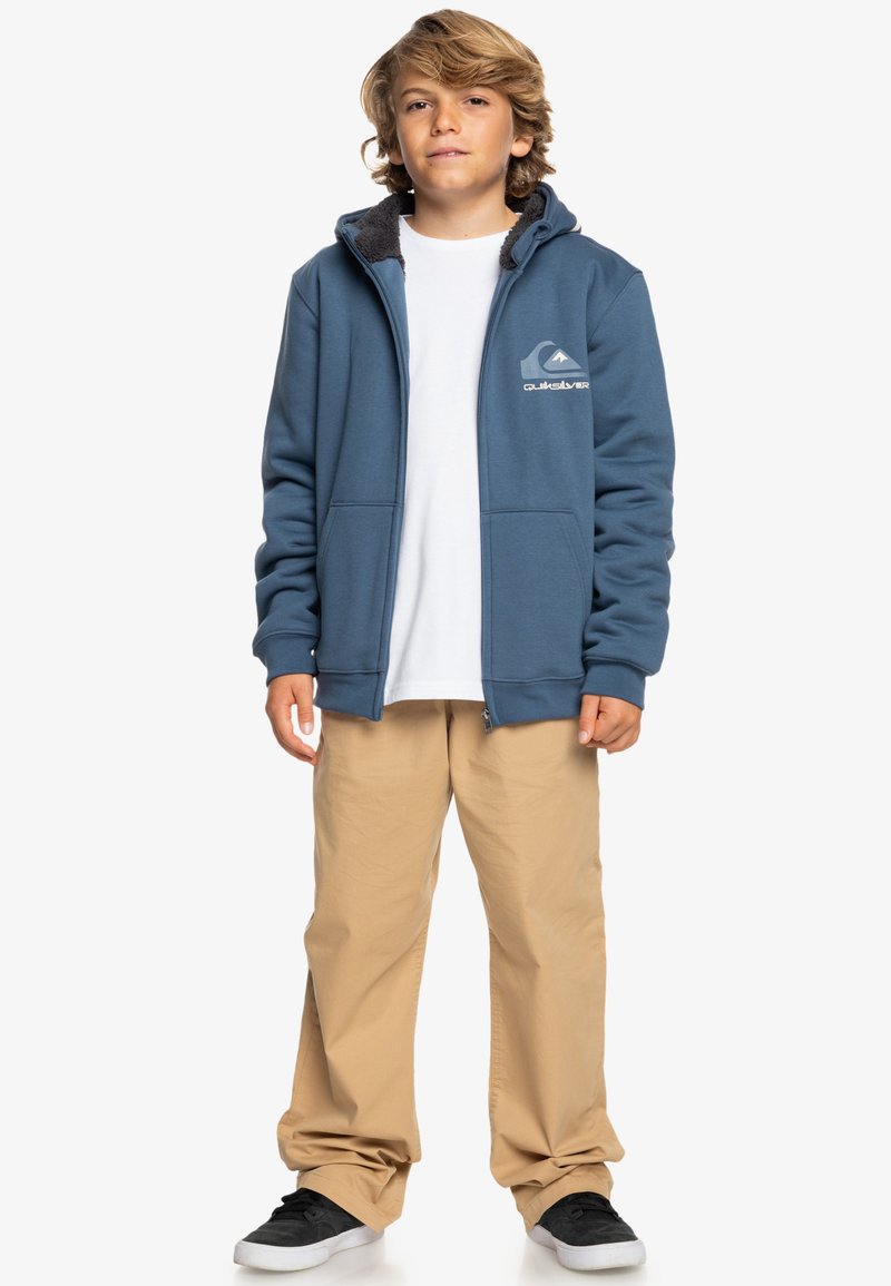 Veste à capuche zippée bleu avec une capuche en fourrure, logo gris sur la poitrine gauche, associée à un pantalon beige et des chaussures noires, t-shirt blanc uni en dessous.