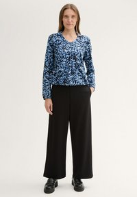 Blaues und schwarzes gemustertes Langarmoberteil mit V-Ausschnitt, kombiniert mit schwarzen weit geschnittenen Hosen und schwarzen Plateauschuhen.