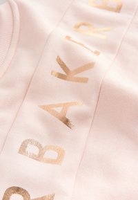 Tissu rose doux avec des lettres dorées brillantes formant une partie du mot "BAKERY" en disposition verticale.