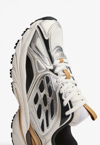 Sneakers z siateczkowym cholewą w kolorze srebrnym i czarnym, białymi sznurowadłami oraz złotymi akcentami; mają teksturowaną podeszwę i wyraźny wycięty design.
