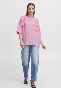 Camicia rosa con colletto alla mandarino, due tasche frontali e maniche corte, abbinata a jeans azzurri larghi e tacchi neri.