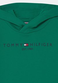 Sudadera verde con bolsillo canguro frontal, con bordado de "TOMMY HILFIGER" en azul marino, con un pequeño logotipo de la bandera y "EST. 1985".