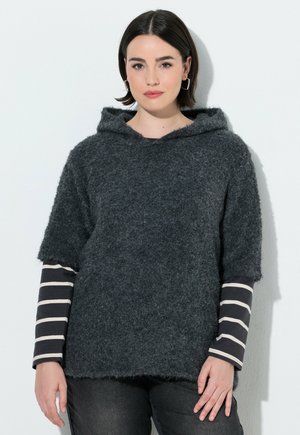 Kvinde med kort mørkt hår iført en mørkegrå, fluffy hættetrøje over en sort-hvid stribet langærmet trøje mod en hvid baggrund.