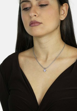 Donna con lunghi capelli scuri che indossa una collana d'argento con un pendente rotondo in pietra preziosa trasparente e un top scuro a scollo a V su sfondo bianco.