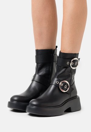 Bottes de cheville en cuir noir avec semelles épaisses et deux larges sangles à boucles argentées, présentées sur des jambes nues sur fond blanc.