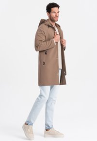Brun hættefrakke med tryknapper, sorte detaljer og en afslappet pasform, parret med lyseblå jeans og beige sneakers.