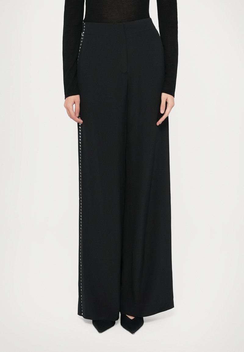 Czarne spodnie o szerokich nogawkach wykonane z gładkiego materiału, z bocznym detalem w białym geometrycznym wzorze. Wysoki pas i fason o pełnej długości.