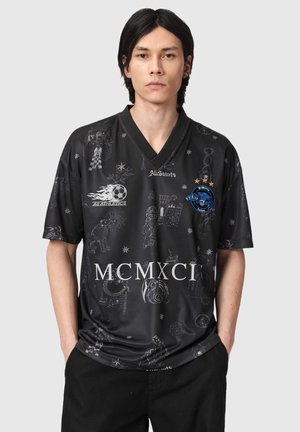 Junger Mann mit schwarzen Haaren, der ein schwarzes AS Athletics-Trikot trägt, das verschiedene graue Grafiken, ein blaues Panther-Emblem und weiße römische Ziffern MCMXCI zeigt.