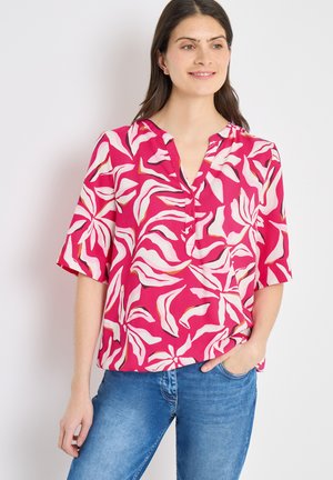 Kvinde iført en skrigpink bluse med store hvide blomsterprint og blå jeans, stående med den ene hånd i lommen og smilende.