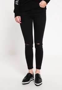 Jeans de mezclilla negros ajustados con detalles desgastados en las rodillas, acompañados de zapatos negros con cordones que tienen una suela de plataforma blanca.