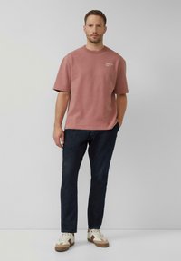 Helles rosa Baumwoll-T-Shirt mit kurzen Ärmeln und Rundhalsausschnitt, versehen mit dezenter weißer Schrift. Kombiniert mit dunklen Jeans und beigen Turnschuhen.