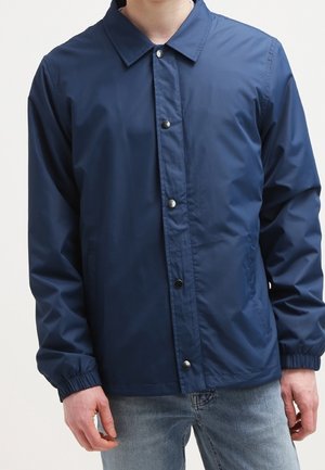 Homme portant une veste boutonnée bleu marine avec des poignets élastiques et un jean bleu clair sur fond uni.