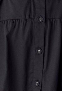 Juodas marškinėlis su mygtukais, turintis penkis mygtukus, raukšlėta tekstūra po krūtinės siūle ir lygus audinio paviršius.