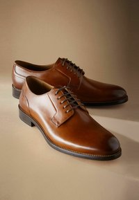 Zapatos de vestir de cuero marrón con un acabado suave, puntera redonda y cordones tradicionales. Presentan un tacón apilado y un detalle de costura en contraste.