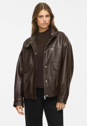 Femme portant une veste bomber en cuir marron foncé par-dessus un pull en maille foncée et un pantalon noir, debout devant un fond clair uni.