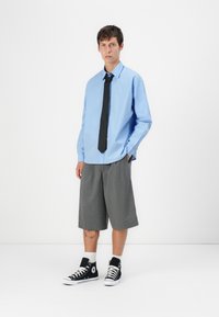 Camicia a maniche lunghe a righe blu chiaro con cravatta nera, abbinata a pantaloni corti grigi plissettati e sneakers alte nere con accenti bianchi.