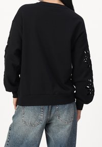 Schwarzer Pullover mit ausgeschnittenen Blumenmustern an den Ärmeln, gerippten Bündchen und Saum. Kombiniert mit hellblauen Jeans.