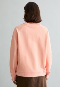 Person mit kurzen braunen Haaren, der einen lockeren, hellrosa Pullover und dunkelbraune Hose trägt, von hinten vor einem einfarbigen Hintergrund gezeigt.