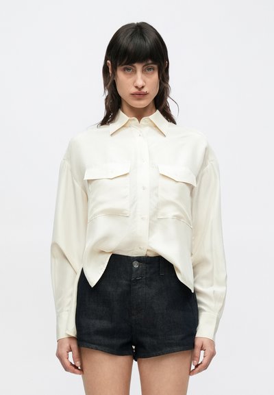 Chemise en satin couleur crème à boutonnage sur le devant, avec deux poches poitrine et manches longues, associée à un short en denim foncé.