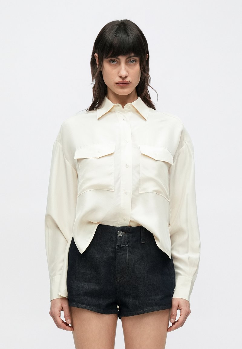 Chemise en satin couleur crème à boutonnage sur le devant, avec deux poches poitrine et manches longues, associée à un short en denim foncé.