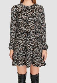 Femme portant une robe à manches longues imprimée léopard avec un ourlet à volants, associée à des bottes noires montantes, sur un fond uni.