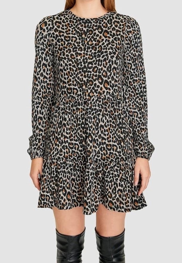 Femme portant une robe à manches longues imprimée léopard avec un ourlet à volants, associée à des bottes noires montantes, sur un fond uni.