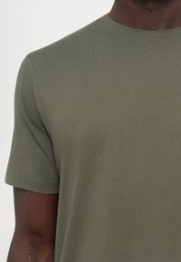 Gros plan d'une personne portant un t-shirt uni vert olive à manches courtes avec un col rond sur fond blanc.