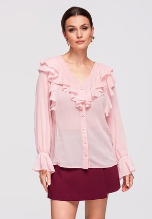 Kobieta ubrana w jasnoróżową falbaniaste bluzkę z długim rękawem i perłowymi guzikami oraz bordową mini spódnicę, stojąca na jednolitym tle.