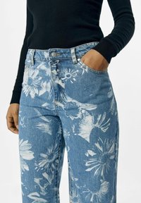 Jeans de mezclilla con estampado floral en azul claro y un patrón de flores blancas. Cuenta con cinco bolsillos, cierre de botón y pasadores para cinturón. El material es algodón.