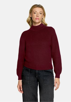 Lyshåret kvinde iført en mørkerød ribbet rullekravesweater og sorte højtaljede jeans mod en ensfarvet hvid baggrund.