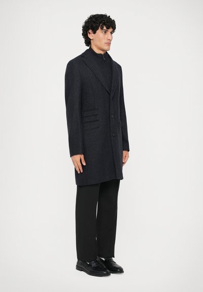 Hombre con abrigo de lana negra sobre suéter con cremallera, pantalones negros y zapatos de cuero negros, de pie contra un fondo claro y sencillo.