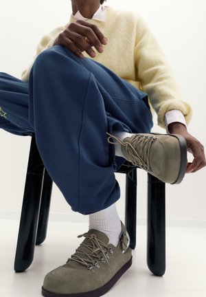 Sárga pulóvert, kék nadrágot csillag részlettel, fehér zoknit és taupe túracipőt viselő személy, aki fekete széken ül.