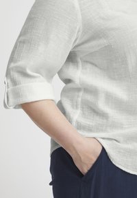 Balta lino marškiniai su trimis ketvirtadaliais rankovėmis ir suktomis rankovėmis, nešiojami su tamsiai mėlynomis kelnėmis, turinčiomis laisvą siluetą ir švelnią tekstūrą.