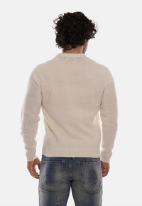 Uomo con capelli ricci e scuri, che indossa un maglione a maglia color crema e jeans blu, con il logo "DS2 Dropseasonz" sulla tasca posteriore, di spalle.