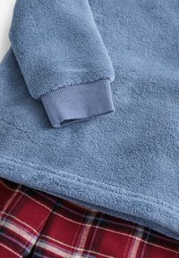 Blaues Fleece-Oberteil mit einer strukturierten Oberfläche und gerippten Bündchen. Darunter zeigt ein rotes Karostoffmuster Farbkontrast und -muster.