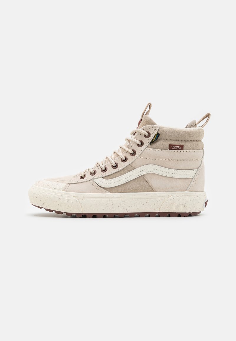 Beige korkeavartiset Vans-tossut, joissa on valkoinen sivuviiva, ruskea pohja, metalliset nyörilinkit, pehmustettu nilkkaremmi ja vetotapit valkoista taustaa vasten.