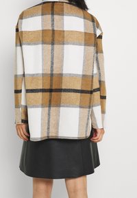 Person klädd i en brun, vit och svart rutig jacka tillsammans med en svart faux-läderkjol, sedd bakifrån mot en enfärgad bakgrund.