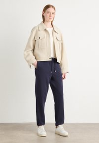 Giacca beige cropped con due tasche frontali, abbinata a pantaloni navy con coulisse e sneakers bianche. Tessuto liscio, design casual.